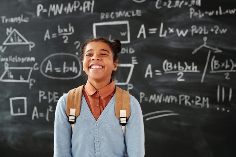 smiling elementary school girl with a backpack, standing in front of a blackboard filled with equations