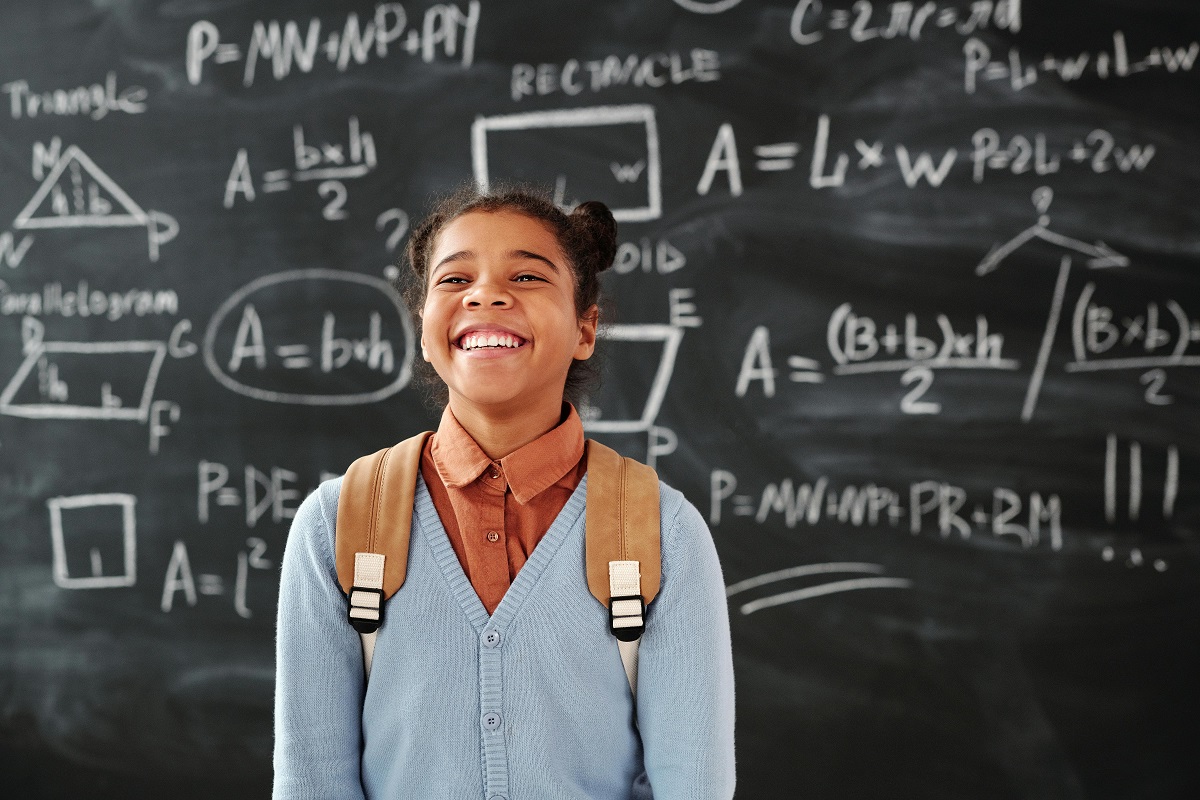 smiling elementary school girl with a backpack, standing in front of a blackboard filled with equations